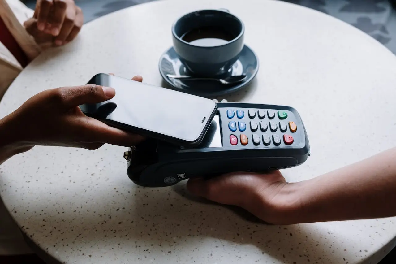 A person using a smartphone to make a contactless payment with a card reader on a cafe table.